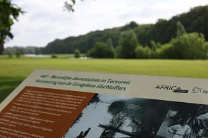 Gedenkplaat voor de slachtoffers van de menselijke zoo van Tervuren in 1897, met de vijvers en het park op de achtergrond. ©Tervuren+