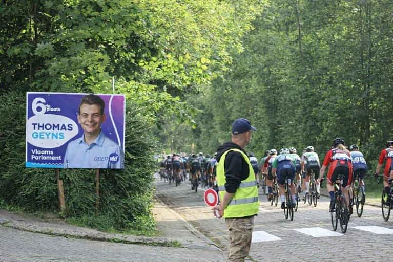 Een groep wielrenners rijdt door Tervuren, met links een verkiezingsbord van Thomas Geyns (Voor Tervuren, Anders/Open VLD).